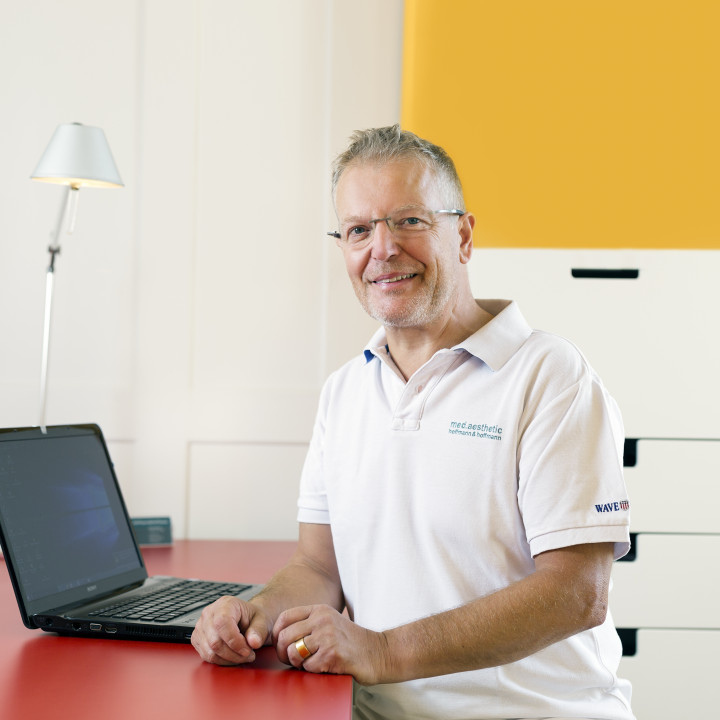 Facharzt Peter Hoffmann sitzt am Schreibtisch vor seinem Laptop und schaut in die Kamera