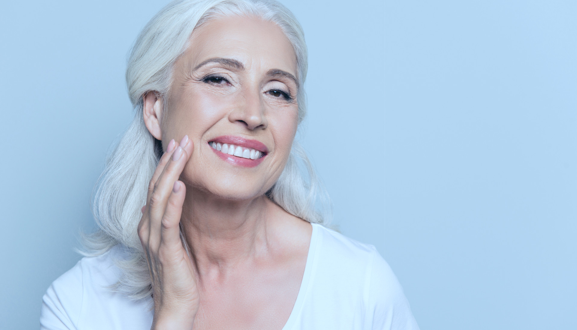 Ältere Frau mit weißen, langen Haaren lächelt in die Kamera und streicht über ihre Wange