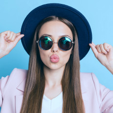 Frau trägt Hut und Sonnenbrille, macht einen Kussmund und hält die Ränder des Hutes fest
