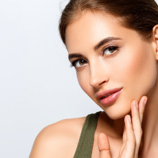 Frau lächelt in die Kamera und fasst sich mit den Fingerspitzen an ihr Gesicht