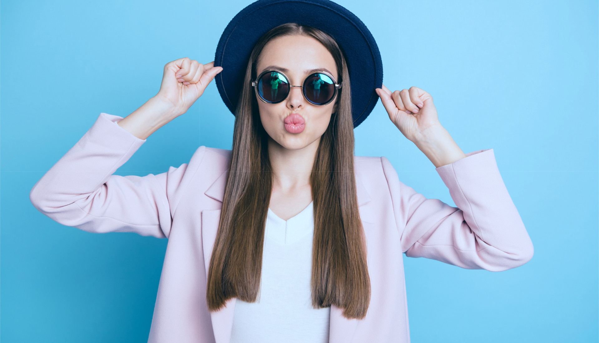 Frau trägt Hut und Sonnenbrille, macht einen Kussmund und hält die Ränder des Hutes fest