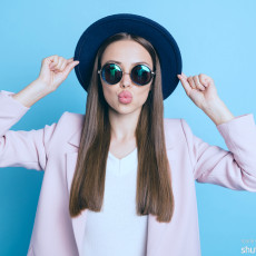 Frau trägt Hut und Sonnenbrille, macht einen Kussmund und hält die Ränder des Hutes fest